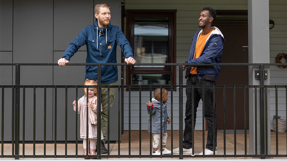 Två pappor står och pratar i en loftgång. Deras barn kikar ut genom räcket. I bakgrunden syns en ingång till en lägenhet och dess förråd. 